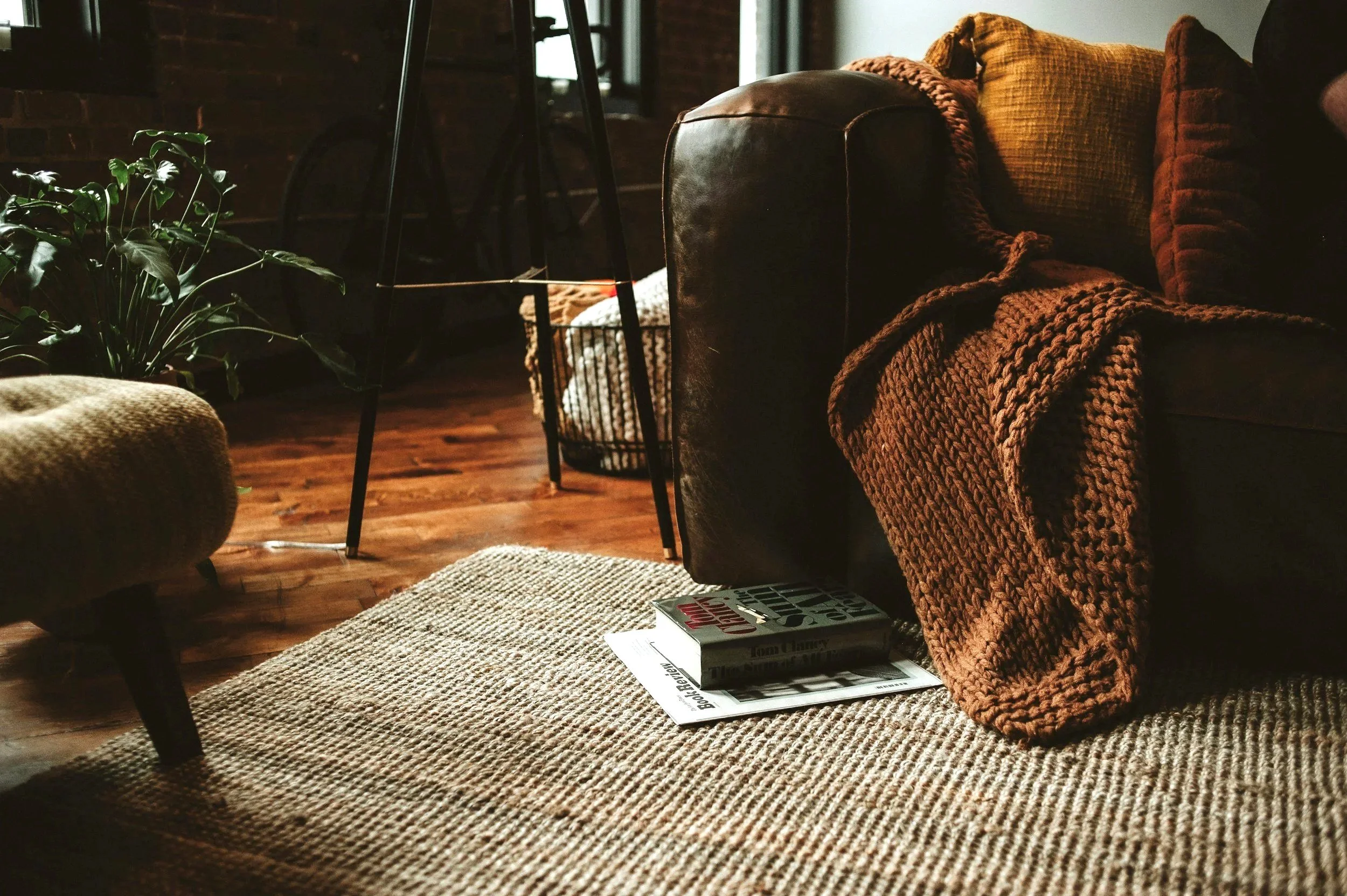 Cozy living room corner with a beige woven rug, a dark leather sofa draped with a chunky knit blanket, two textured pillows, a potted plant with dark green leaves, a stack of books, and a magazine on the rug. In the background, there's a black metal stool and a laundry basket, with natural light illuminating the space.