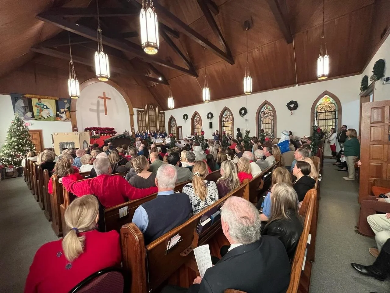 A Christmas service inside a church filled with a large congregation of people seated on wooden pews, decorated with Christmas wreaths and ornaments, and a Christmas tree on the left side. The church features stained glass windows, wooden paneling on