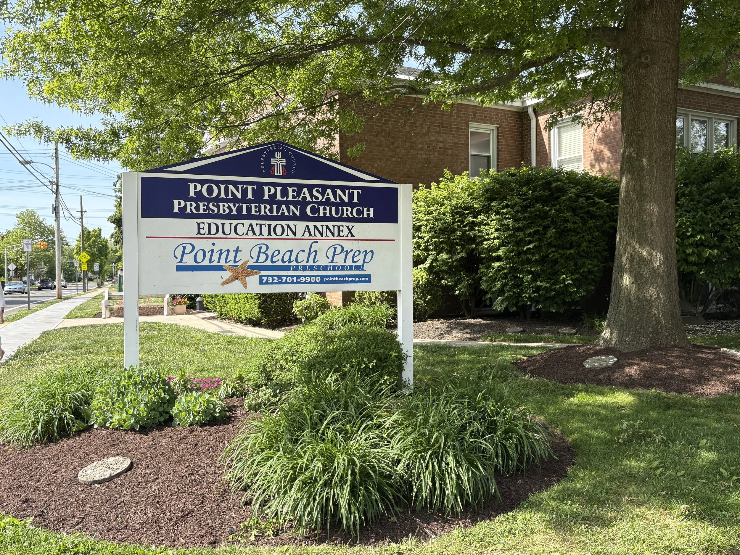 A sign for Point Pleasant Presbyterian Church's education annex and preschool, with trees and bushes in front, and a building in the background, along a street with traffic and power lines.