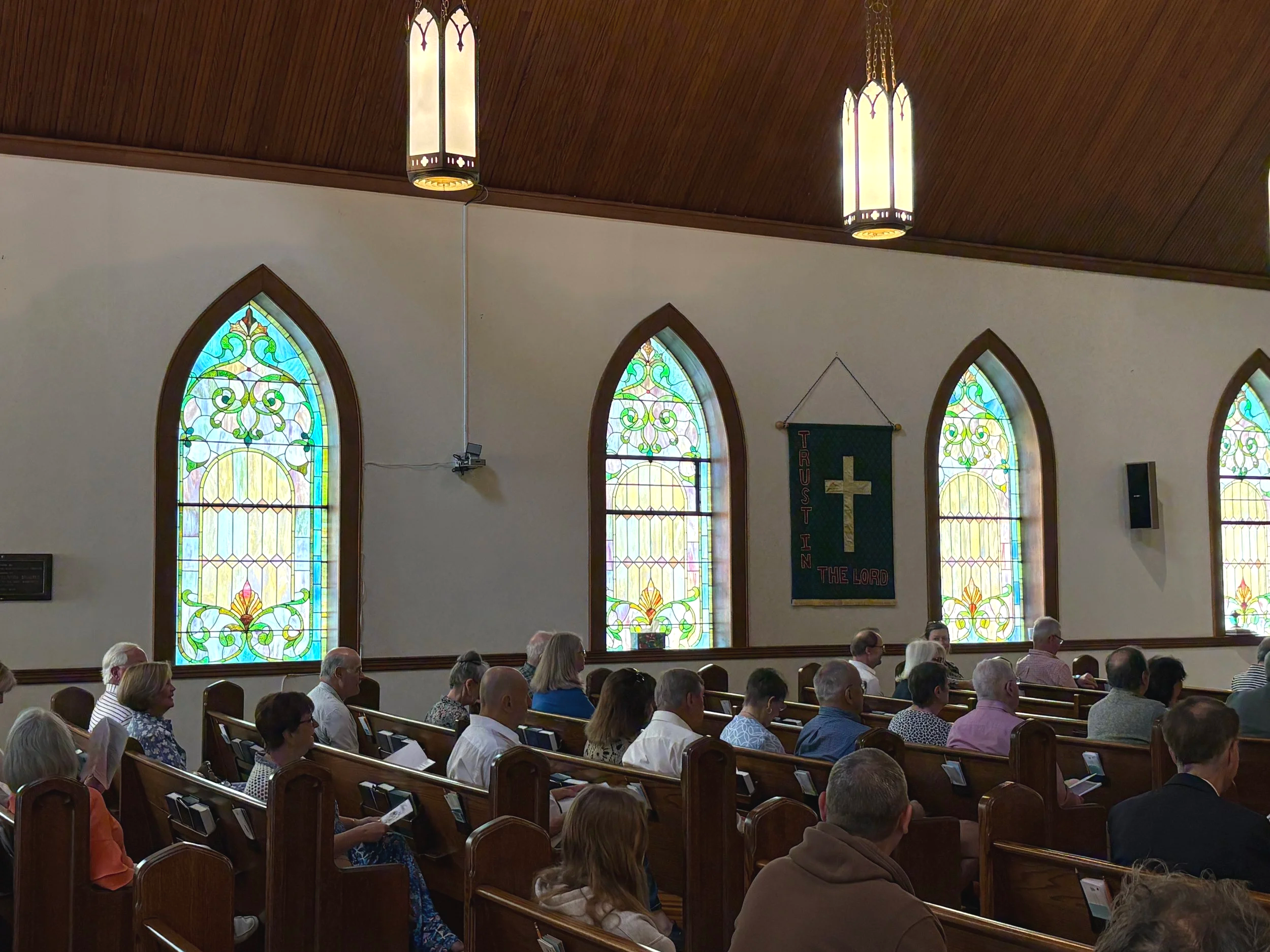 Inside a church with stained glass windows, people seated in pews, and hanging light fixtures.
