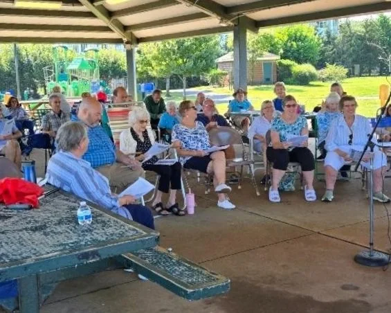 Group of elderly people sitting on chairs under a pavilion, listening to someone speak or present outside on a sunny day.