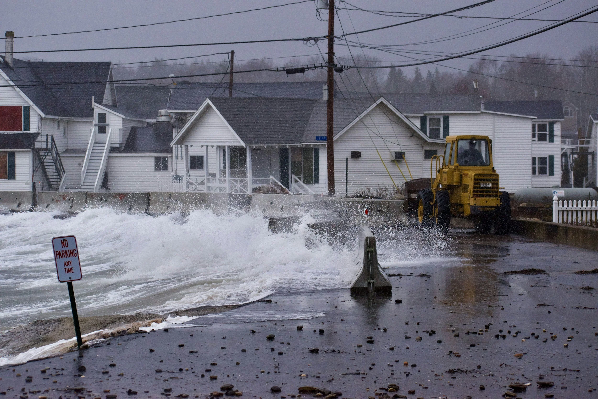 Flooded street with a yellow construction vehicle and a 'No Parking' sign knocked over. Water is splashing as waves crash against the sidewalk. Houses and utility poles line the background on a rainy, overcast day.
