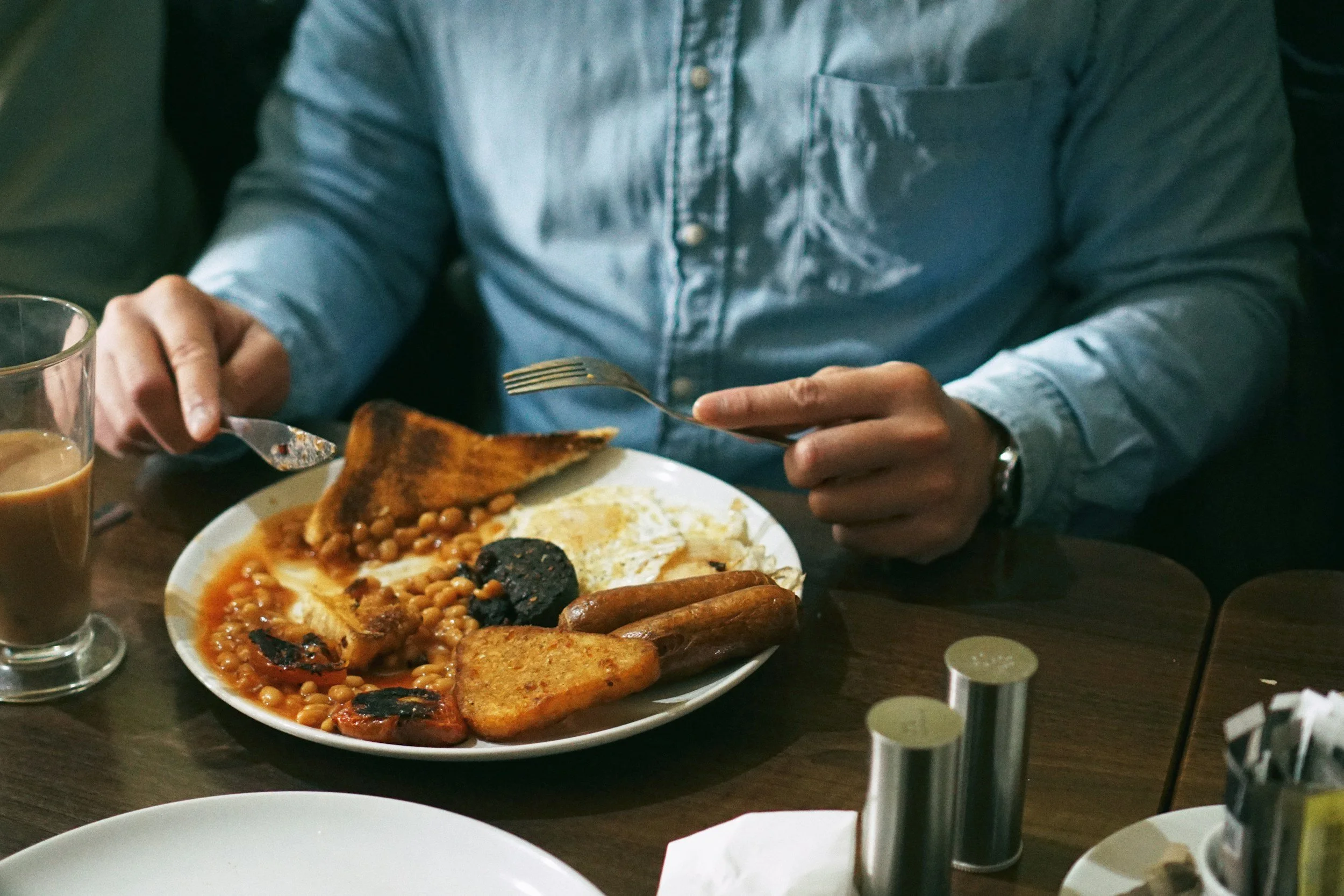 Person wearing a light blue denim shirt eating a plate of breakfast food including baked beans, toast, eggs, and sausage, with a glass of tea or coffee on the table.