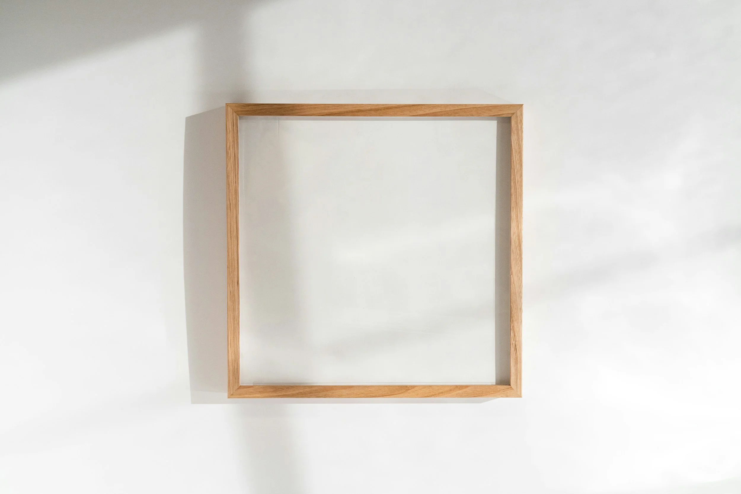 Empty rectangular wooden picture frame leaning against a white wall with shadows.