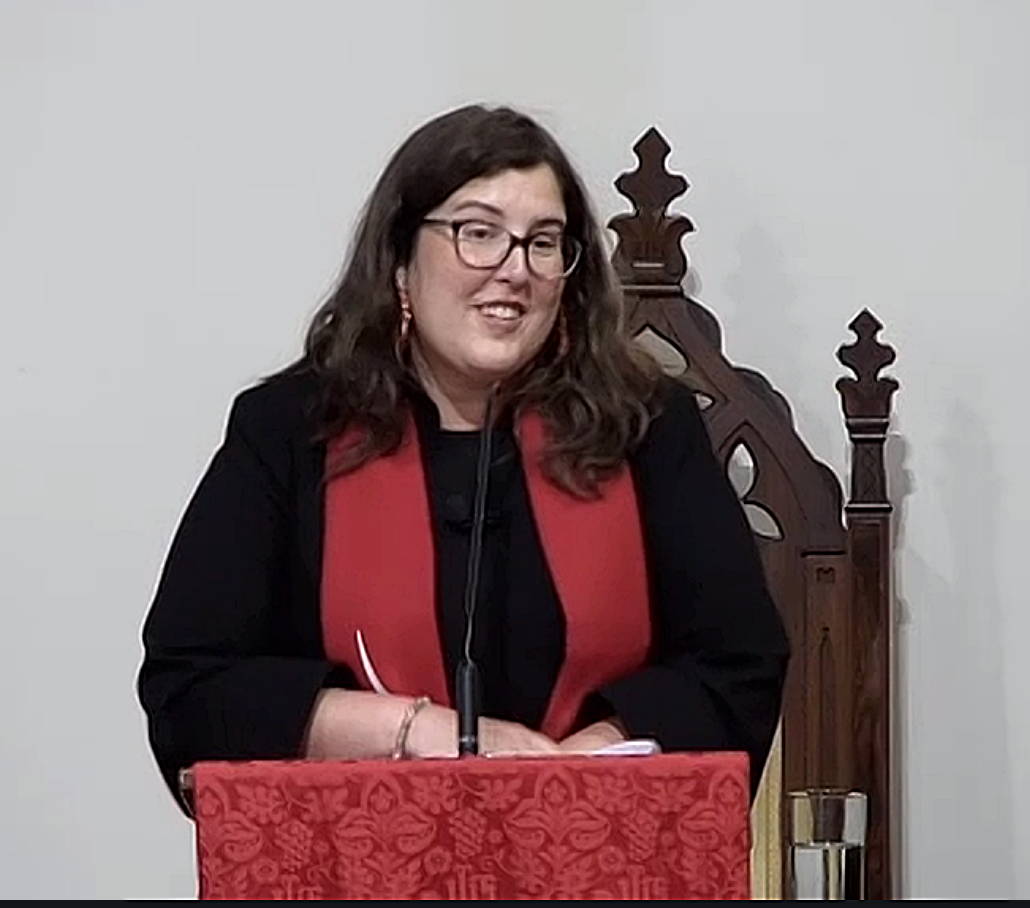 A woman with glasses and long dark hair smiling while standing behind a red decorated podium, wearing a black outfit with a red scarf, in front of a white wall with a wooden chair behind her.
