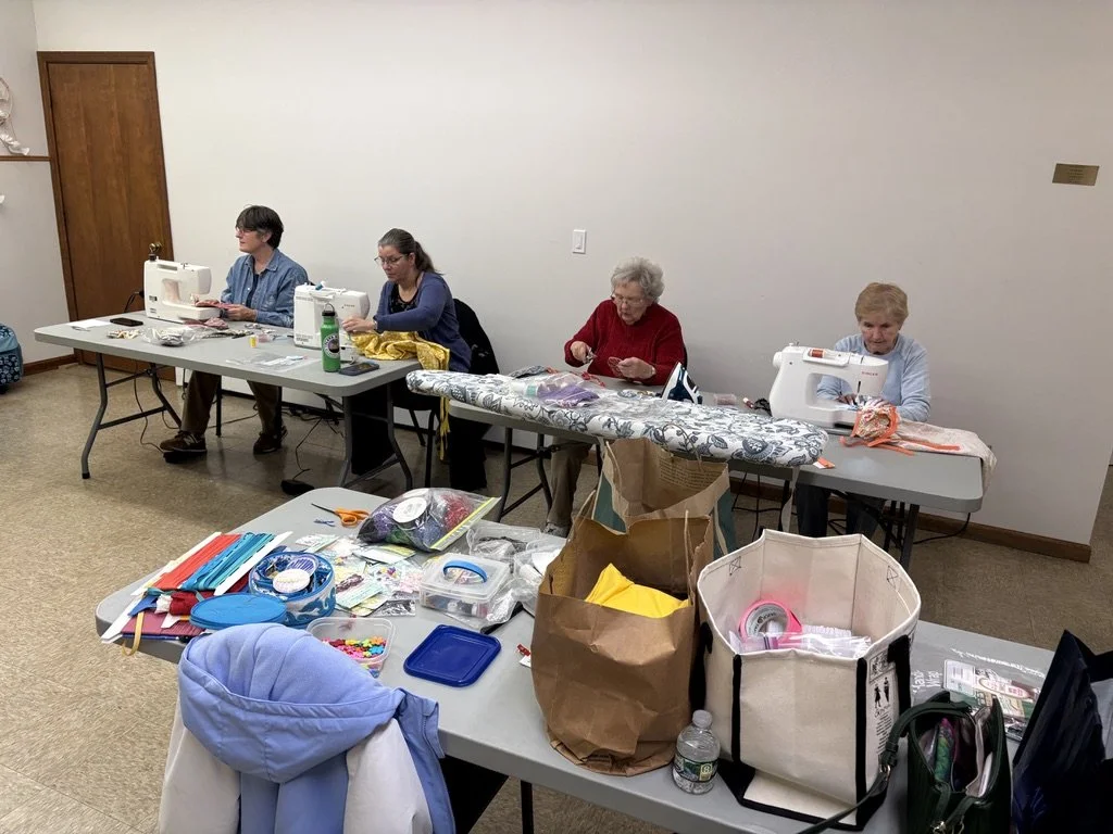 Four women sewing at a long table in a room with crafting supplies and fabric, with a second table in the foreground holding colorful craft items and bags.