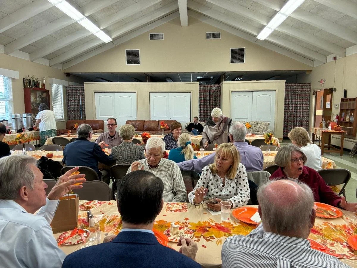 A group of people gathered around tables for a social event in a decorated room with fall-themed tablecloths and decorations.