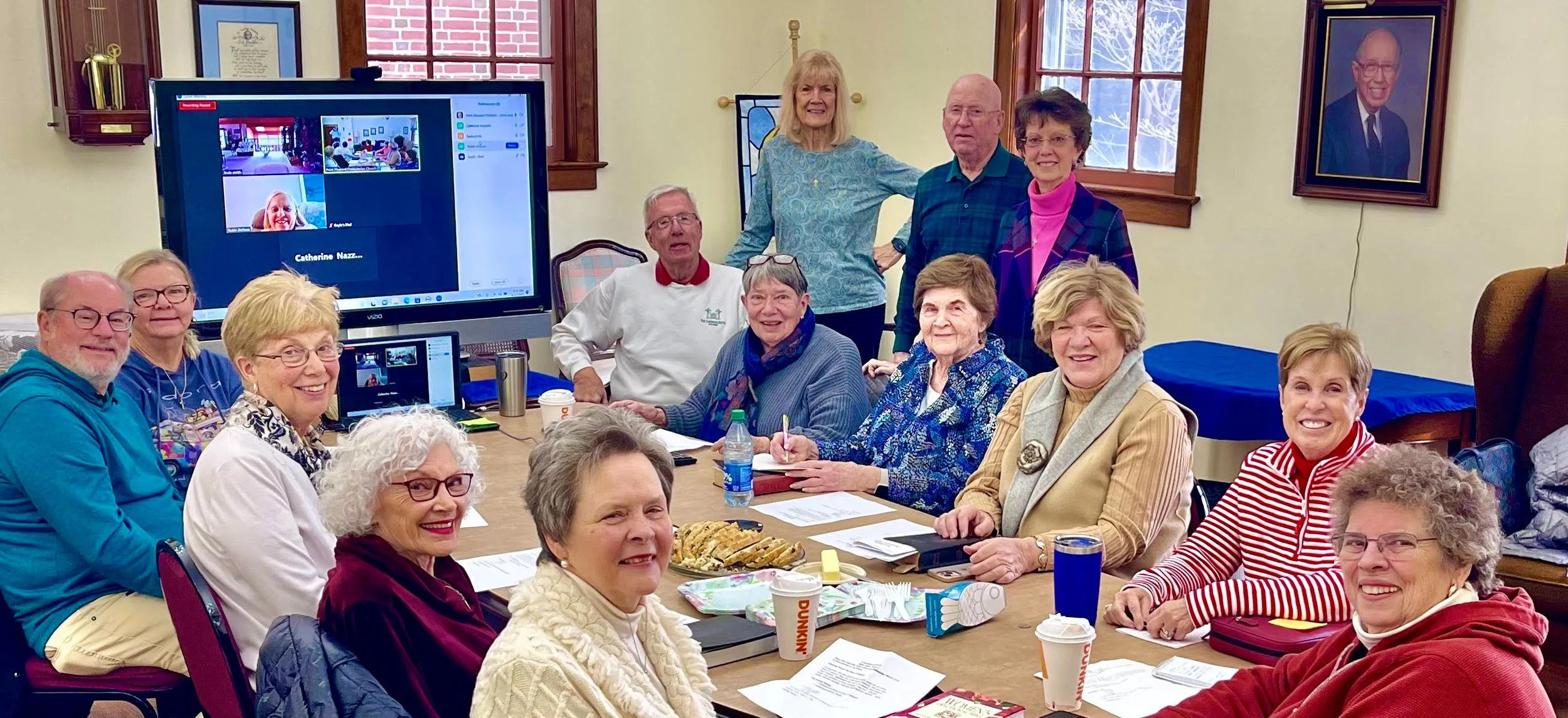 A group of older adults gathered around a conference table in a room, smiling at the camera. Some are seated, while others stand behind. There are papers, notebooks, coffee cups, and snacks on the table. A large TV screen displays a video conference with a few participants, and a framed portrait hangs on the wall.