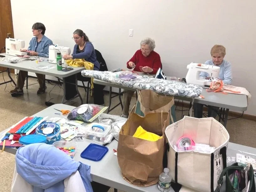 Four women sitting at tables using sewing machines with various sewing supplies on the tables, including fabric, scissors, and containers.