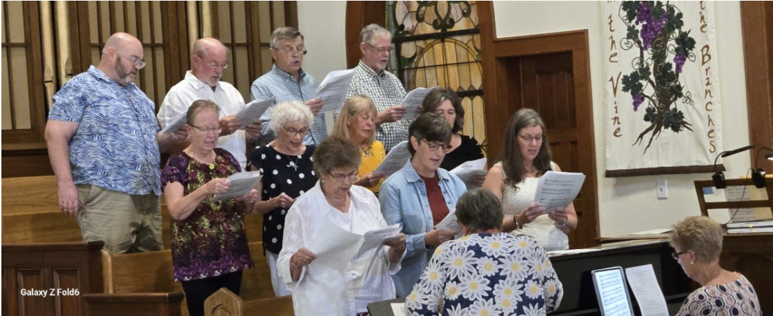 A diverse group of men and women singing in a choir church setting, with some holding sheet music, led by a pianist, in front of a banner with grapes and the words 'the vine' and 'the branch'.