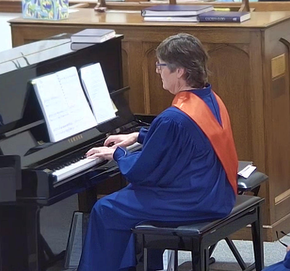 A woman in a blue graduation gown and orange sash playing a black upright piano with sheet music in a church or formal setting.