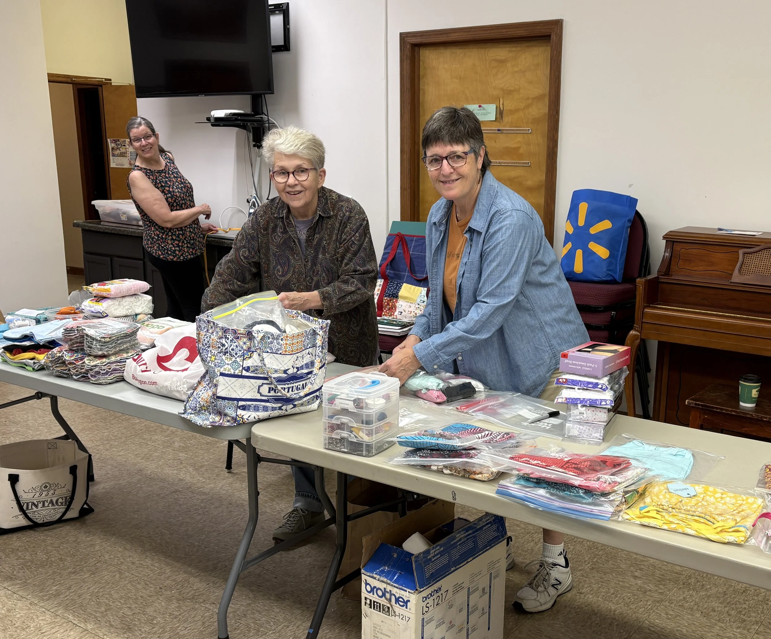 Four women in a room with tables full of fabric, sewing supplies, and a printed box. Two women are sitting at the table, one standing in the background near a counter. They are smiling or looking at the camera.
