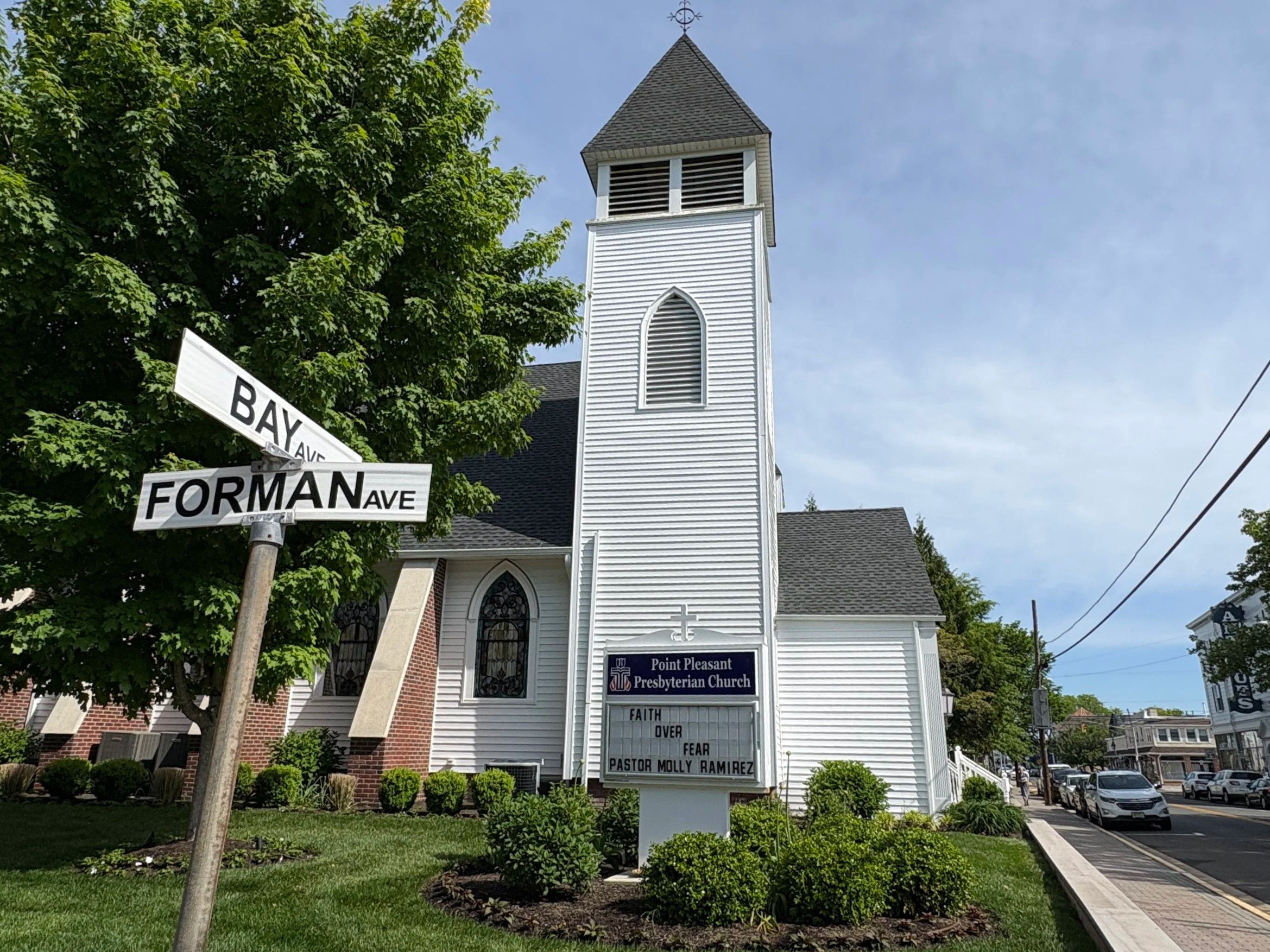 A white church building with a pointed steeple and a sign that reads Point Pleasant Presbyterian Church. There are street signs for Bay Avenue and Forman Avenue at the corner and green trees around the church. The sky is blue with a few clouds.
