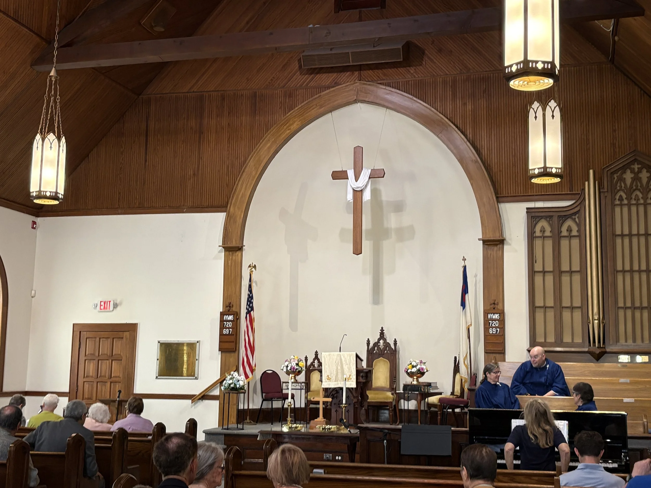 Interior of a church with a wooden ceiling, hanging light fixtures, a cross on the far wall, and people gathered inside facing the altar area.