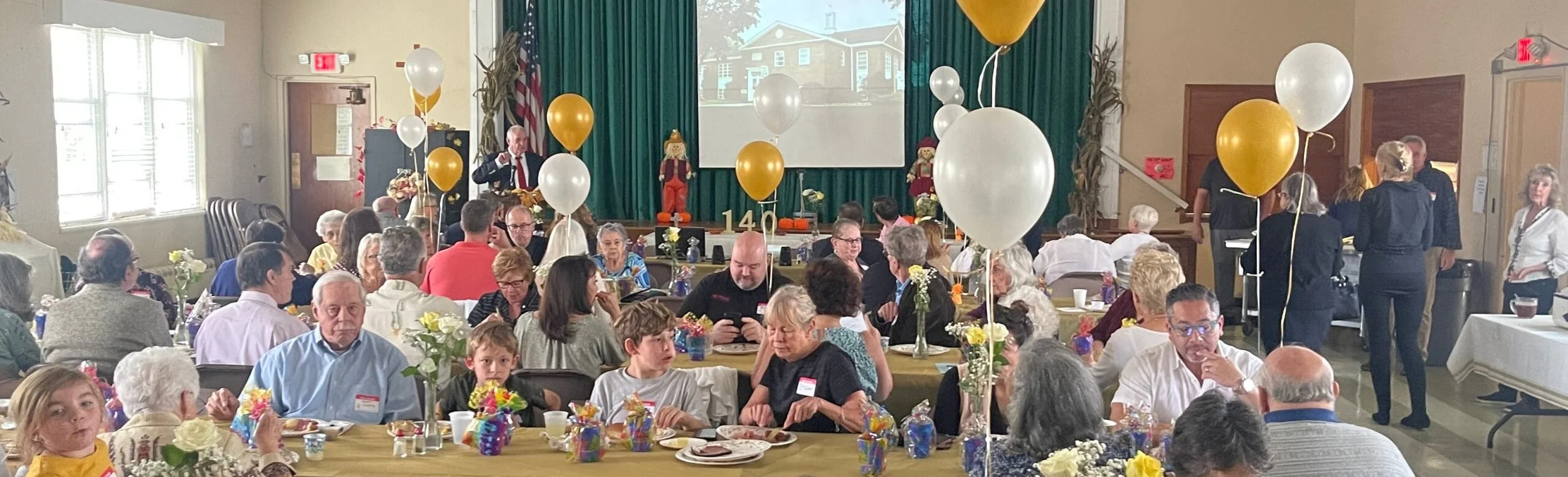 People gathered in a decorated hall for a celebration with balloons, flowers, and food, while a speaker addresses the crowd on stage.
