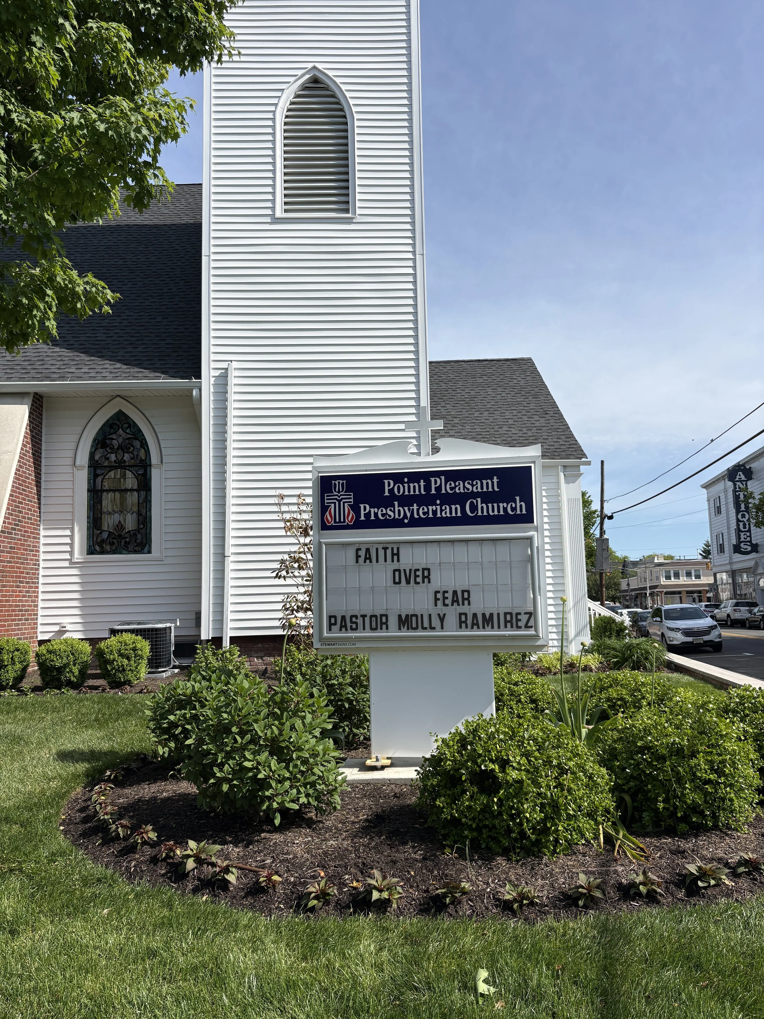 Point Pleasant Presbyterian Church sign with religious message, surrounded by well-maintained garden and trees, near a church building with white siding and arched windows.