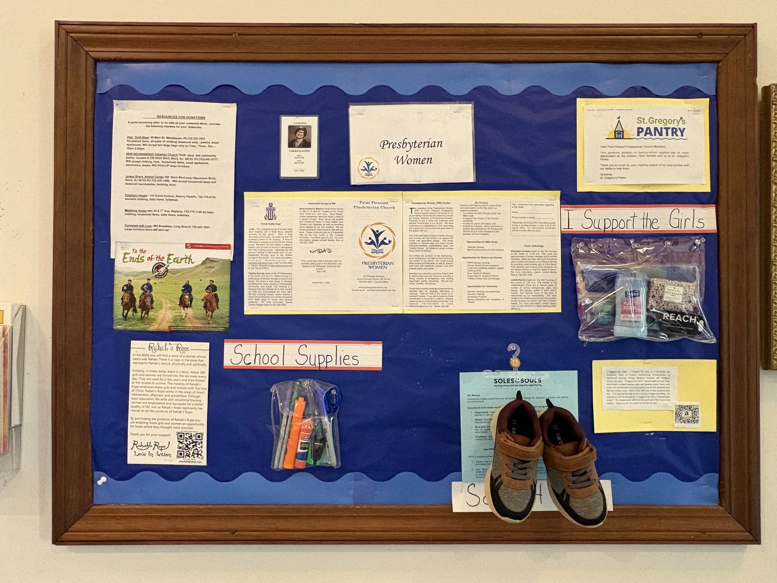 A bulletin board with blue backing and a wooden frame, displaying flyers, notes, and a pair of children's shoes hanging at the bottom. One flyer features a group of people riding horses across a landscape, and handwritten signs read "School Supplies"