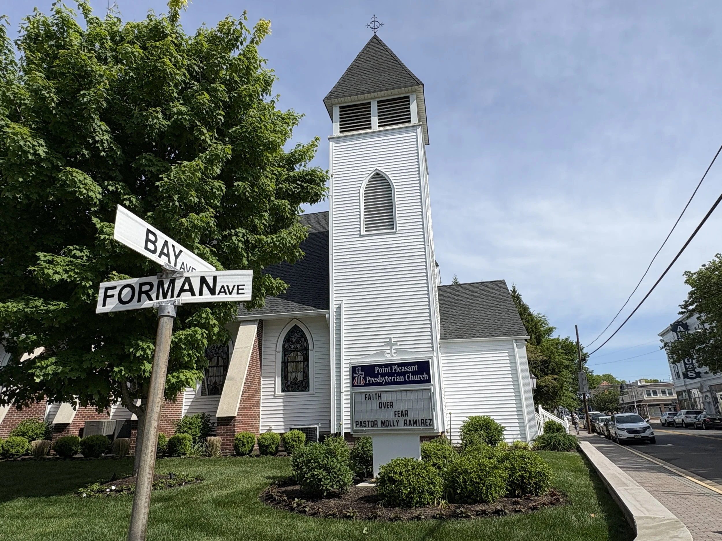 The image features the exterior of Point Pleasant Presbyterian Church, a white wooden church with a tall steeple. There are street signs for Bay Avenue and Forman Avenue on a pole in the foreground, a large leafy green tree, and a well-maintained gar