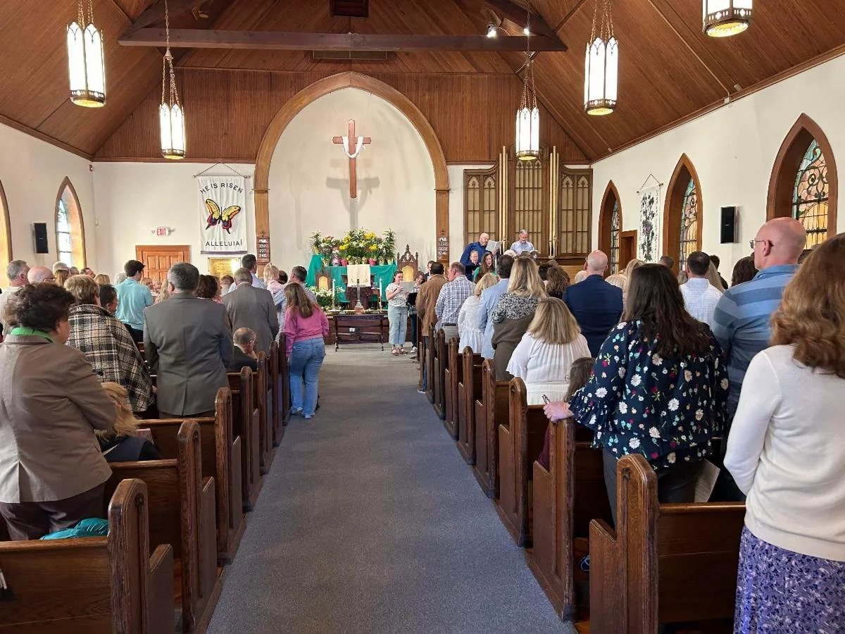 People gathered inside a church, standing during a service. The church has wooden pews, arched windows, and an altar decorated with flowers and religious items. A crucifix is visible on the wall behind the altar.