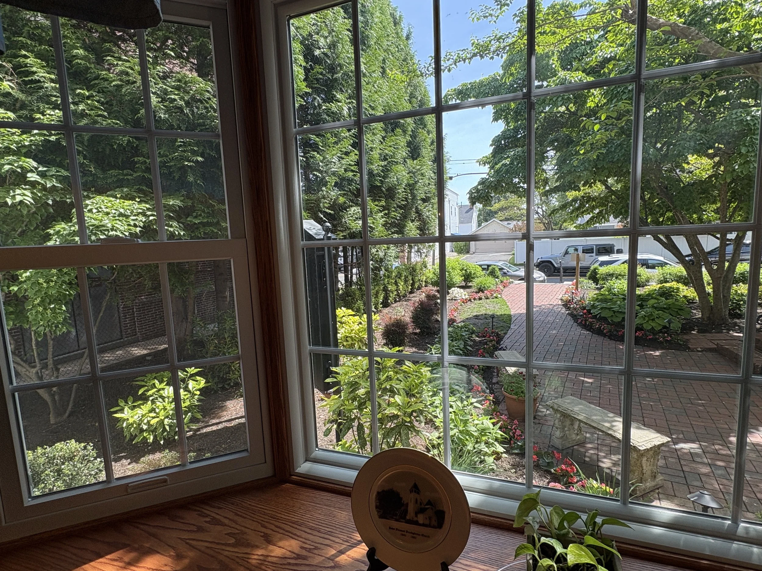 View through a home window showing a sunny garden with lush green trees, a brick pathway, and parked cars in the neighborhood.