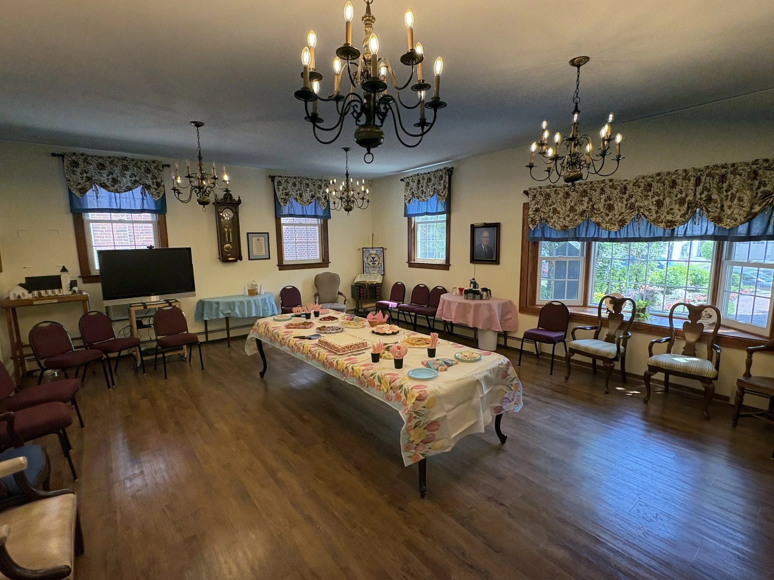 A decorated banquet or gathering room with multiple chandeliers hanging from the ceiling and windows with floral valances. There's a long table with a colorful tablecloth, desserts, and pink napkins in small cups. Several chairs are arranged around t