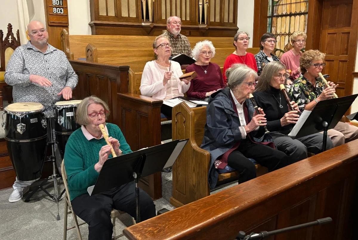 A group of people singing and playing musical instruments in a church or community hall. Some are playing recorders, and there are percussion instruments in the background.