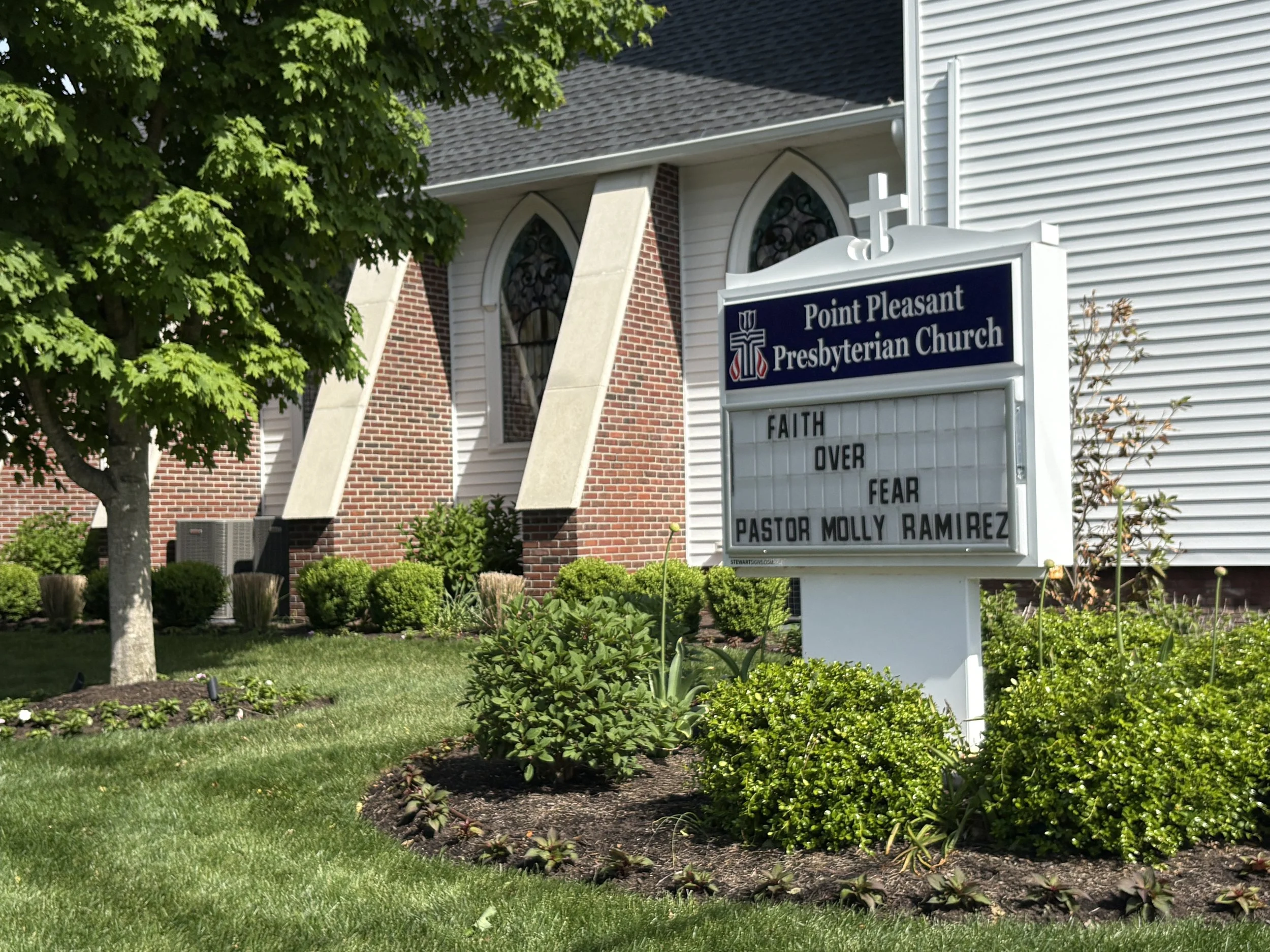 Sign in front of Point Pleasant Presbyterian Church, surrounded by trees and landscaped bushes, with stained glass windows visible on the church building.