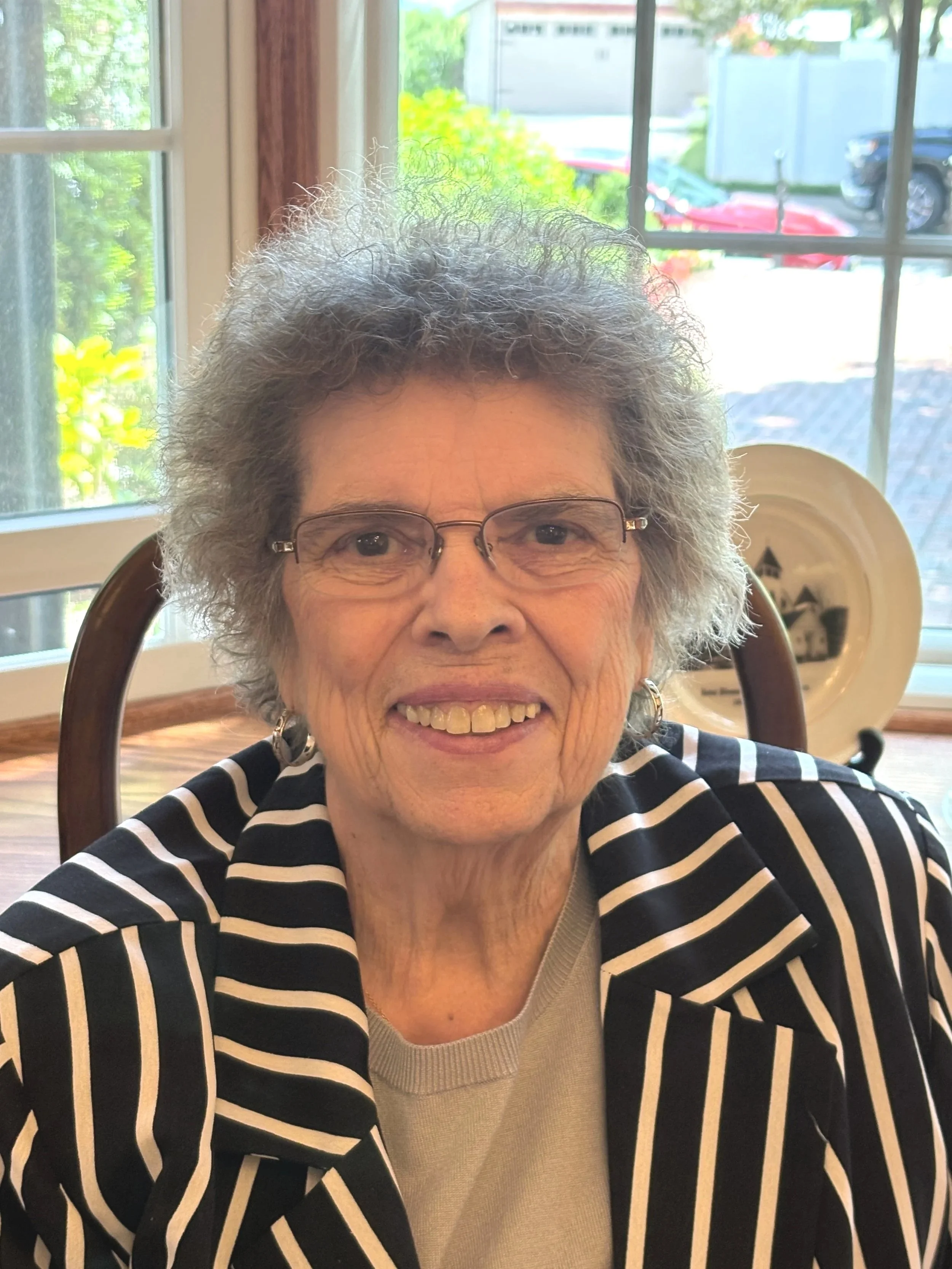 A smiling elderly woman with glasses and curly gray hair sitting indoors at a wooden table near a window, wearing a black and white striped blazer over a beige top.
