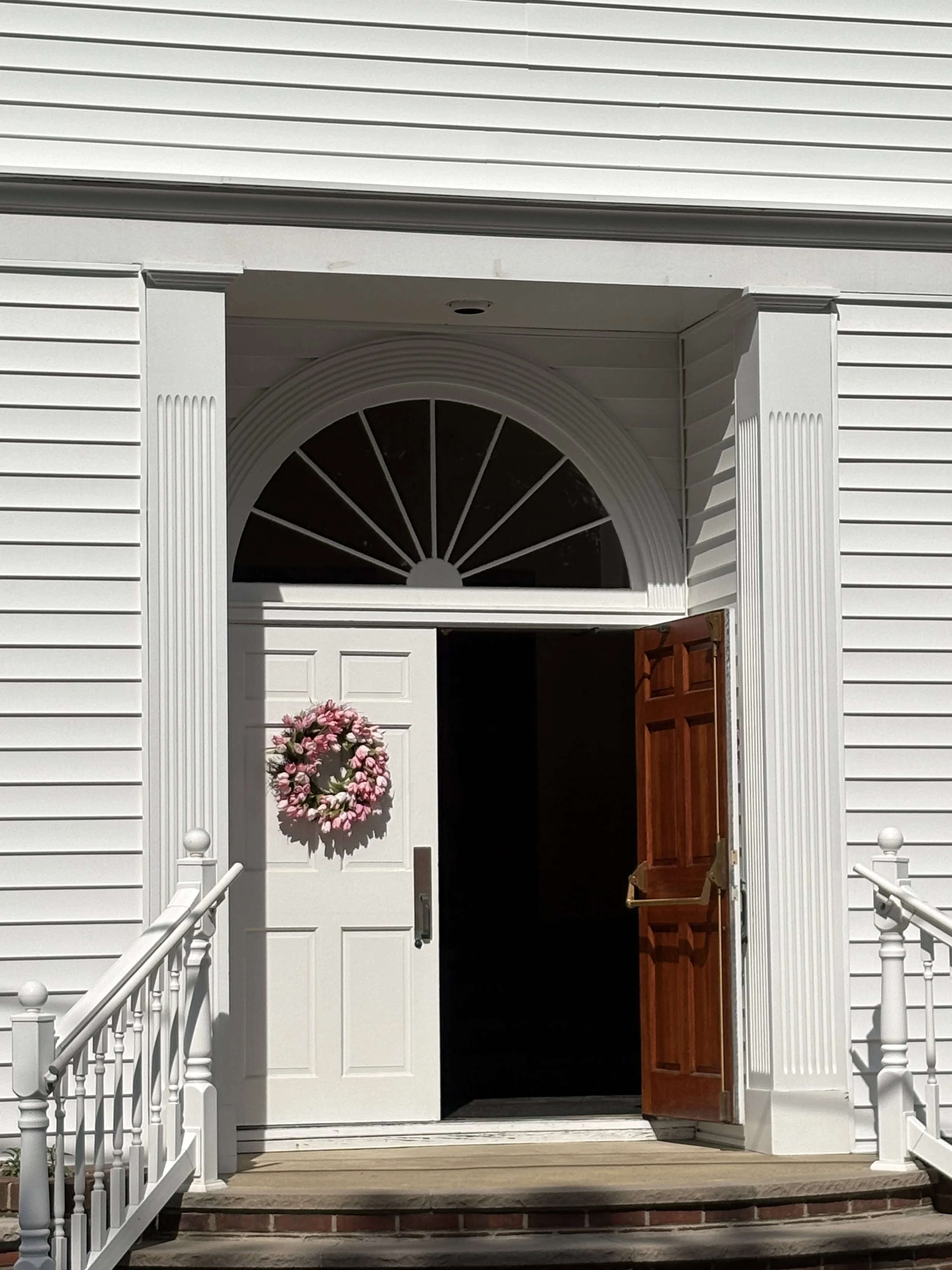 White house with a brown front door, a wreath hanging on the white door, and white porch railing and stairs.