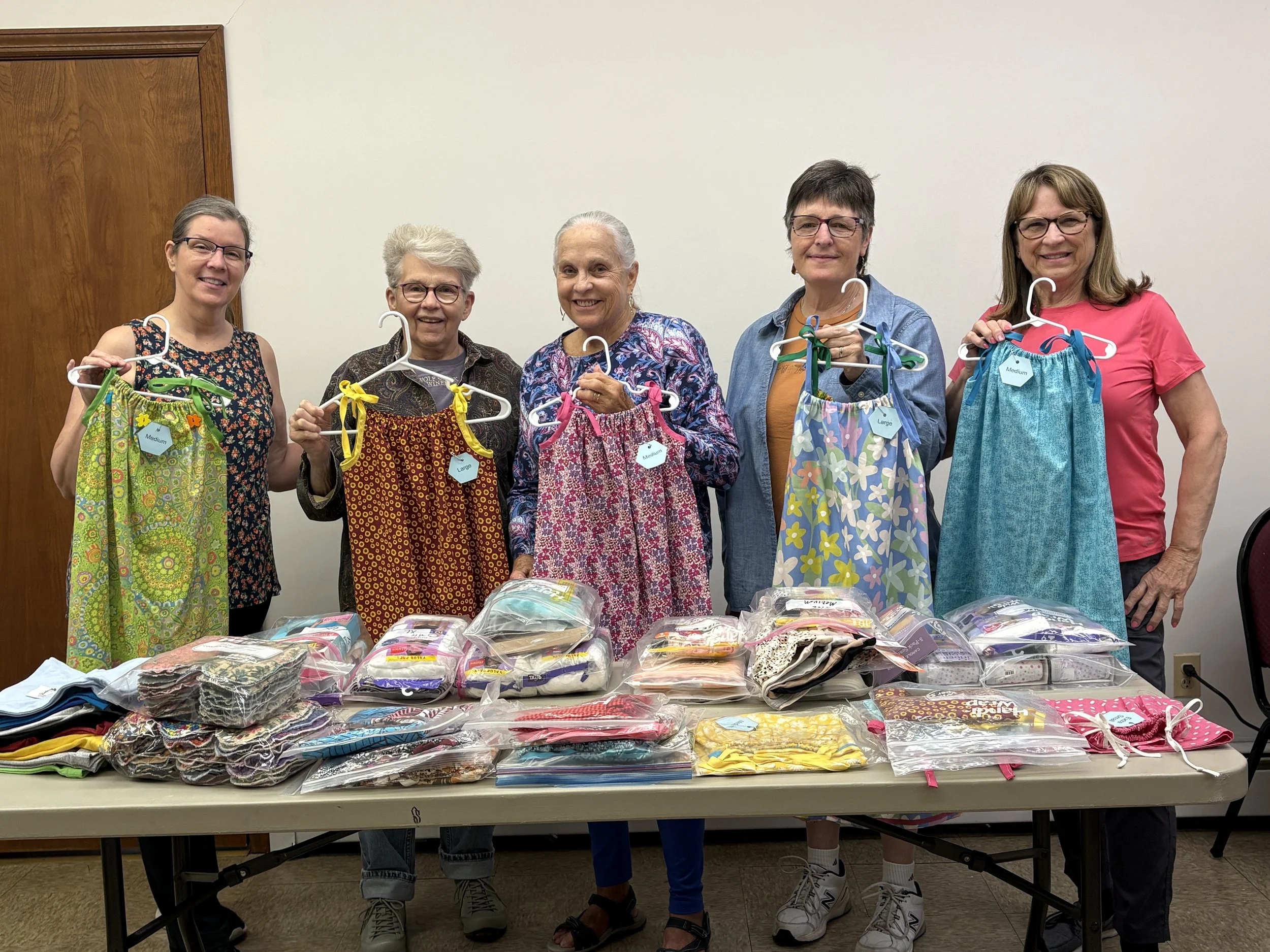 Five women standing behind a table with various folded dresses. They are holding dresses on hangers, some labeled with sizes like medium and large. The women are smiling and appear to be at a clothing donation or distribution event indoors.