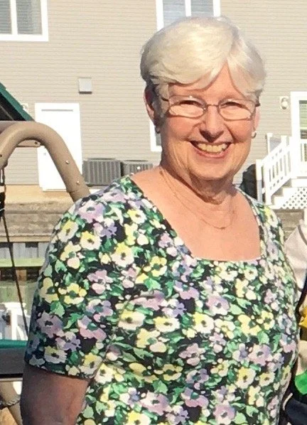 Smiling elderly woman with short white hair and glasses, wearing a colorful floral shirt, standing outdoors in front of a house and a boat.