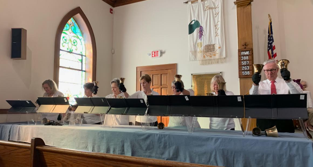 Seven people in a church, each holding a brass bell and wearing headphones, standing behind a long table with black music stands, with a stained glass window and informational hymn number board in the background.