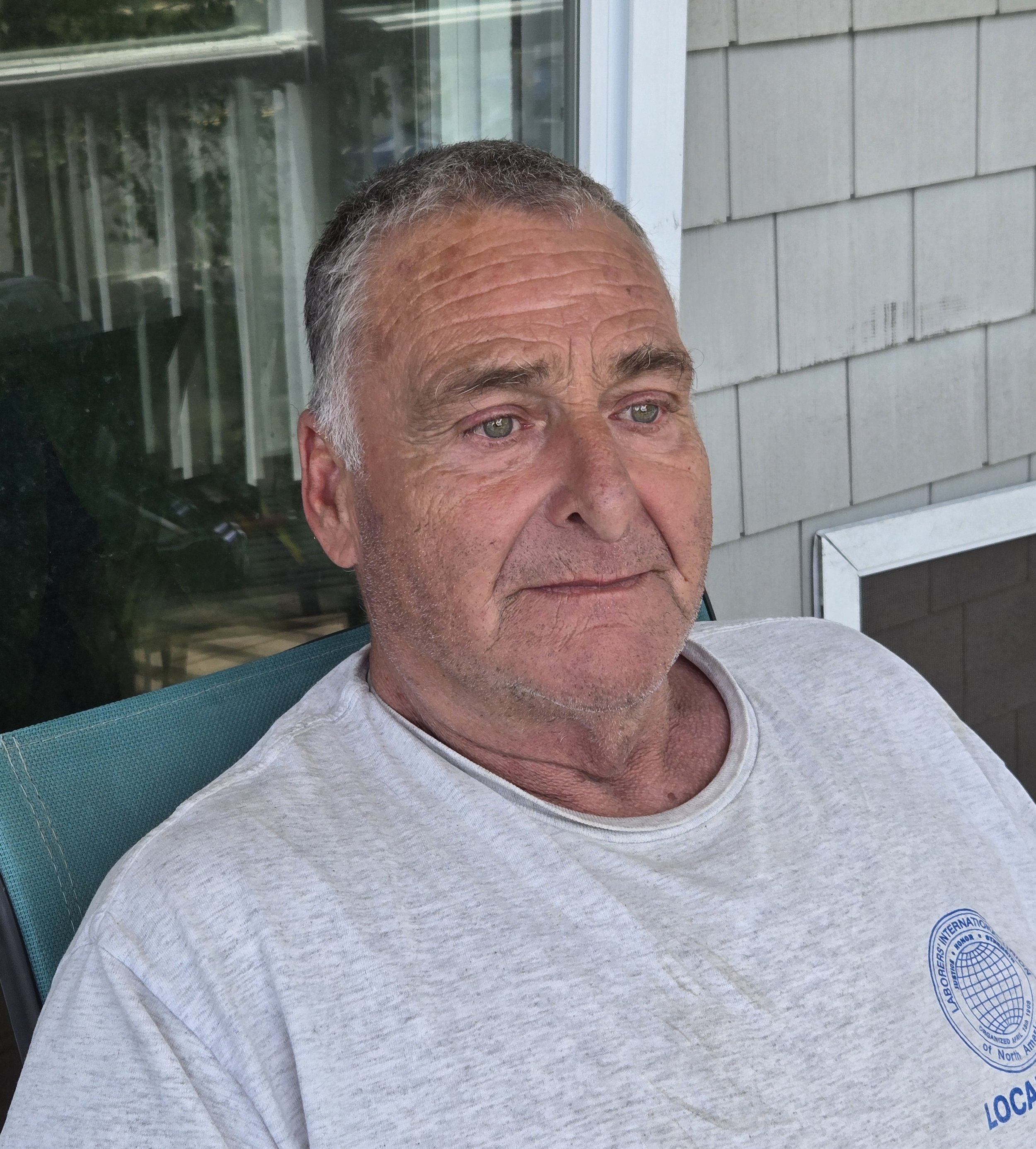 A close-up of a middle-aged man with graying hair, sitting outdoors on a teal chair, wearing a light gray shirt with a logo on the sleeve, in front of a house's exterior wall with light-colored shingles and a window with curtains.