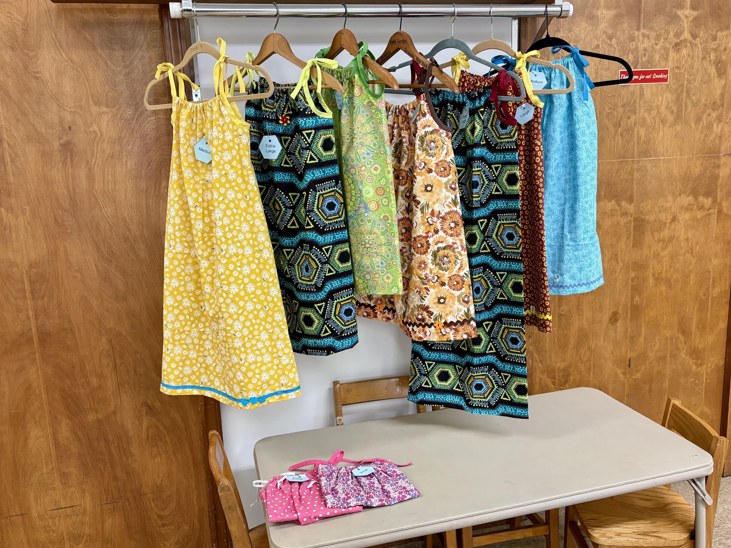 Display of children's clothing on hangers and folded items on a table, including colorful dresses and sets with tags indicating size.