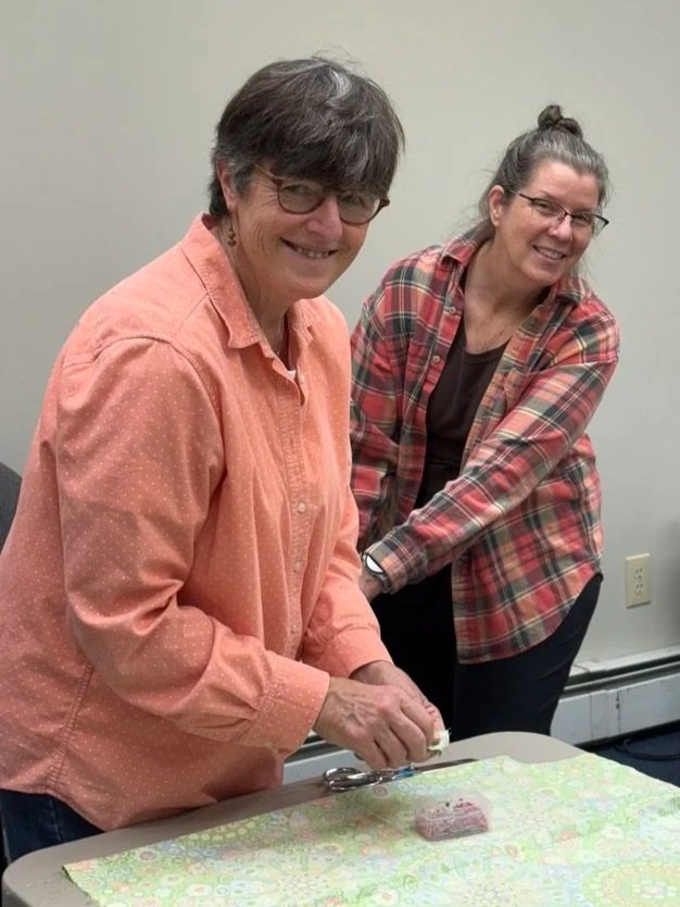 Two women smiling, one with short hair, glasses, and a peach-colored shirt, cutting a small cake on a table covered with pastel-colored wrapping paper, in a room with plain walls.