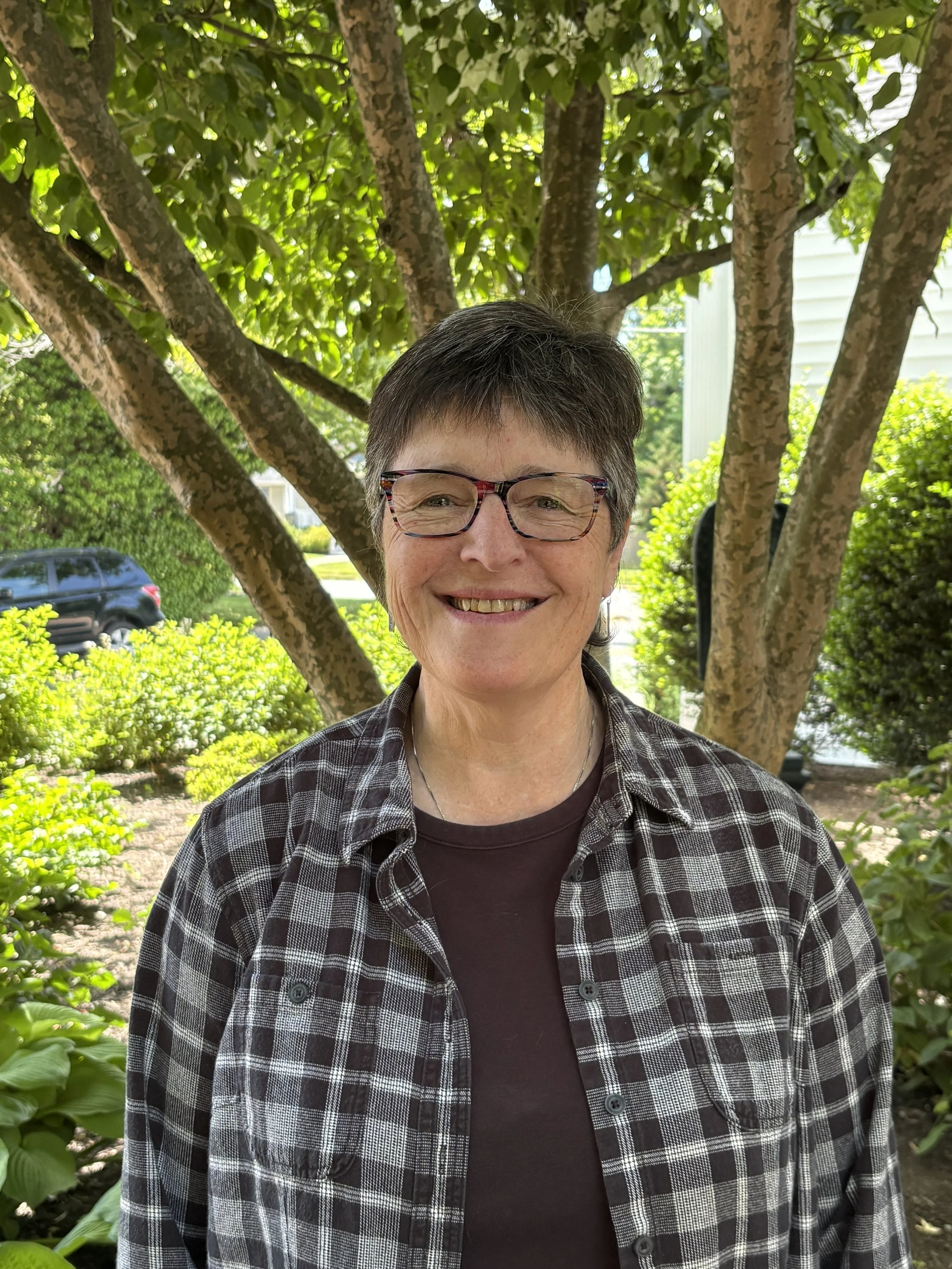 A smiling woman with short gray hair, glasses, and a plaid shirt, standing in front of a tree with green leaves and bushes in a garden or park setting.