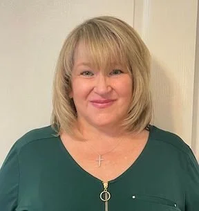A woman with blonde hair, wearing a green top with a gold zipper and a cross necklace, smiling at the camera.