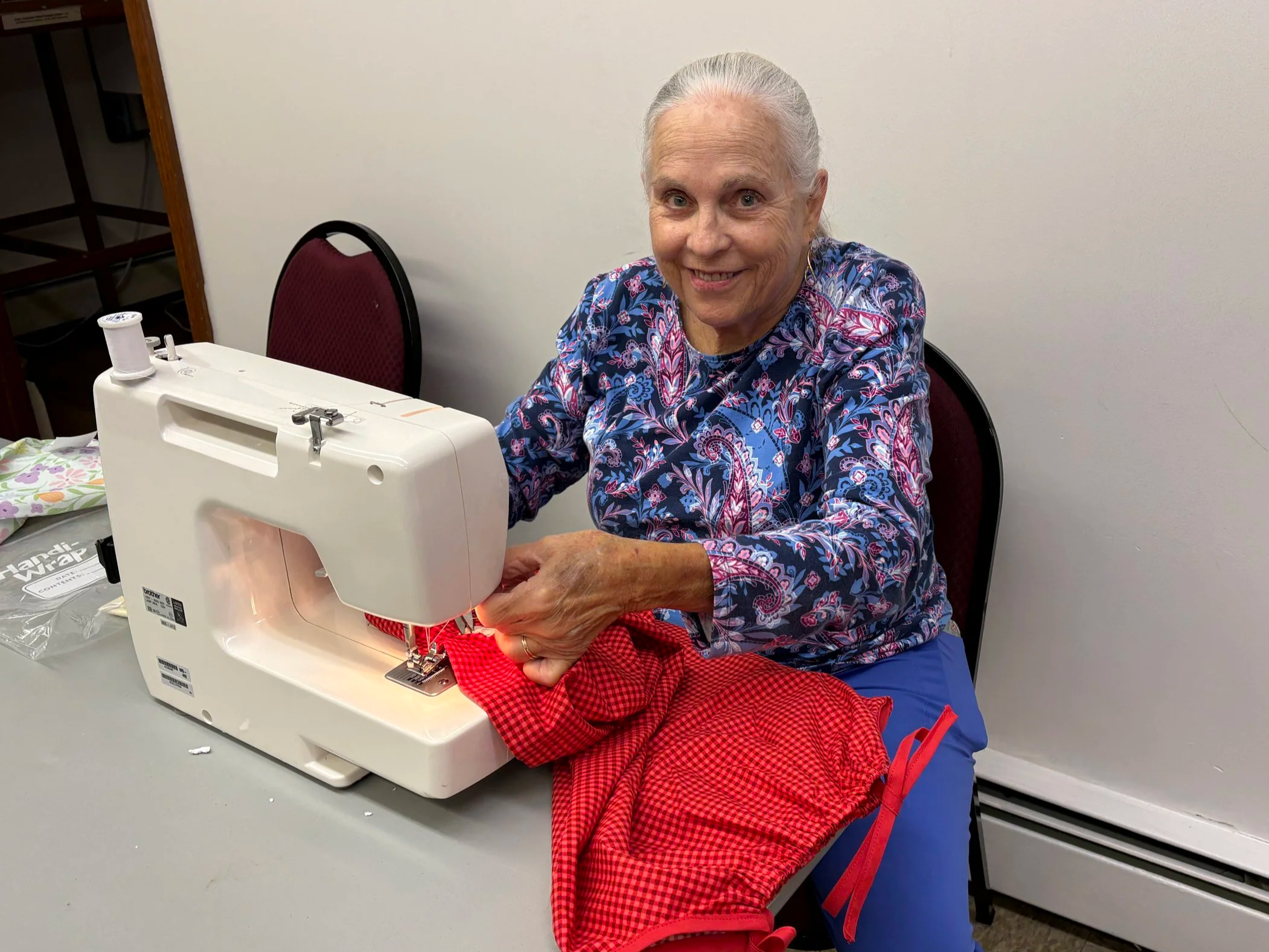 An elderly woman with gray hair sewing fabric on a sewing machine at a table in a room with plain white walls.