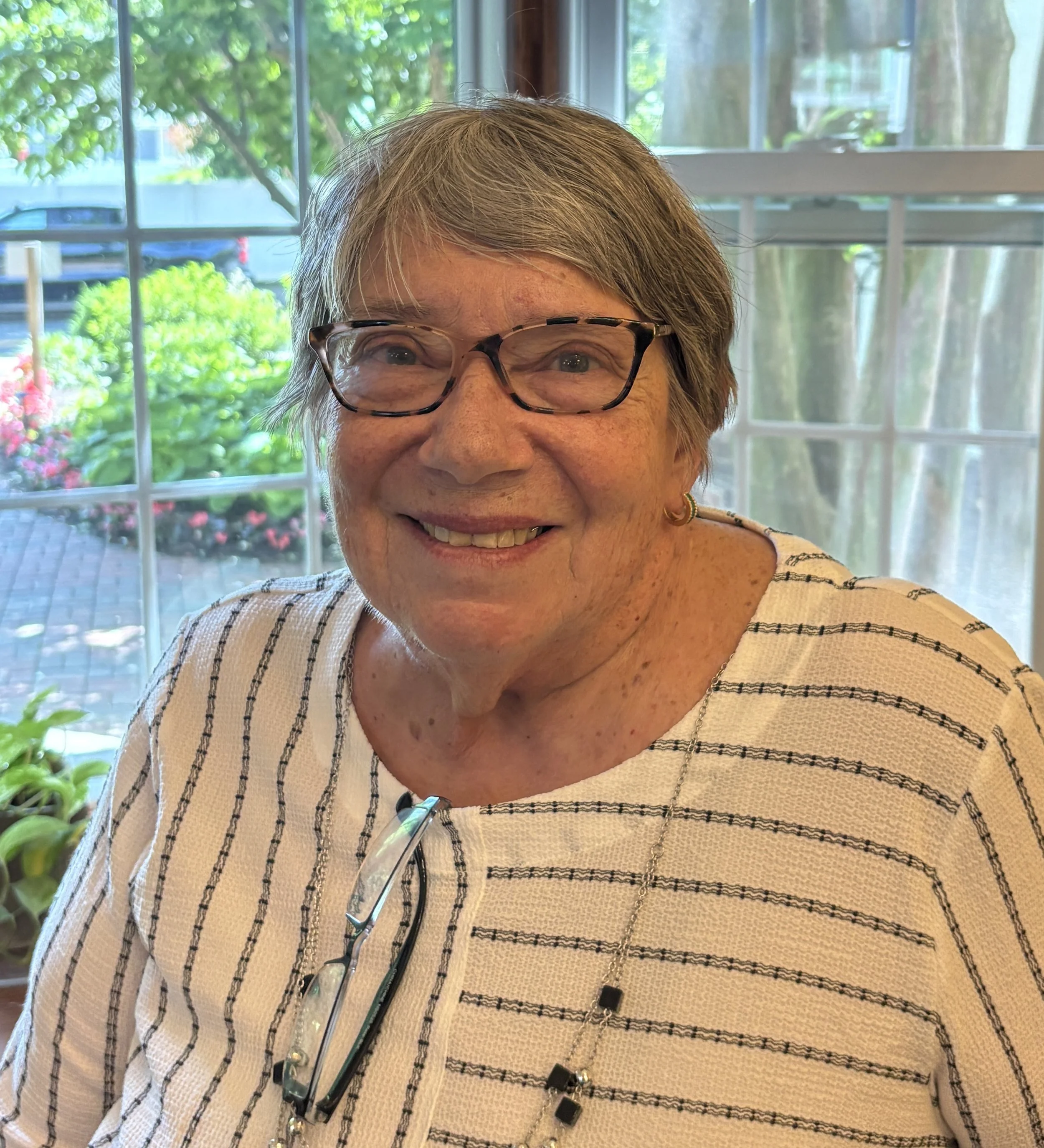 Close-up of a middle-aged woman with short gray hair wearing glasses, smiling, sitting indoors near a window with a view of trees and plants outside.
