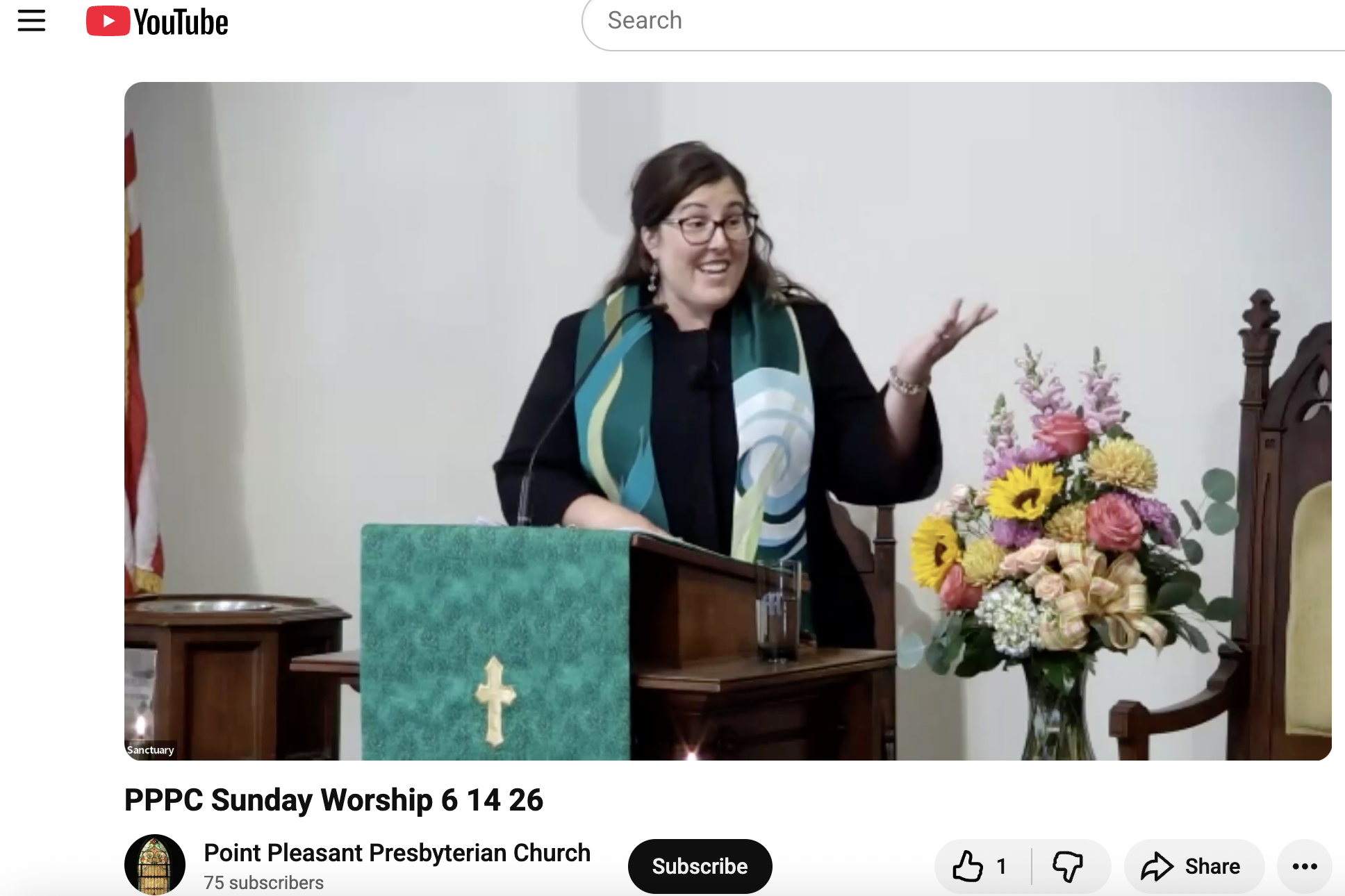 A woman with glasses and dark hair, wearing a colorful scarf, is speaking at a church service. She is standing behind a wooden pulpit decorated with a green cloth featuring a gold cross. To her right is a bouquet of pink, yellow, and purple flowers on a stand. An American flag is visible in the background, along with white walls and wooden church furniture.