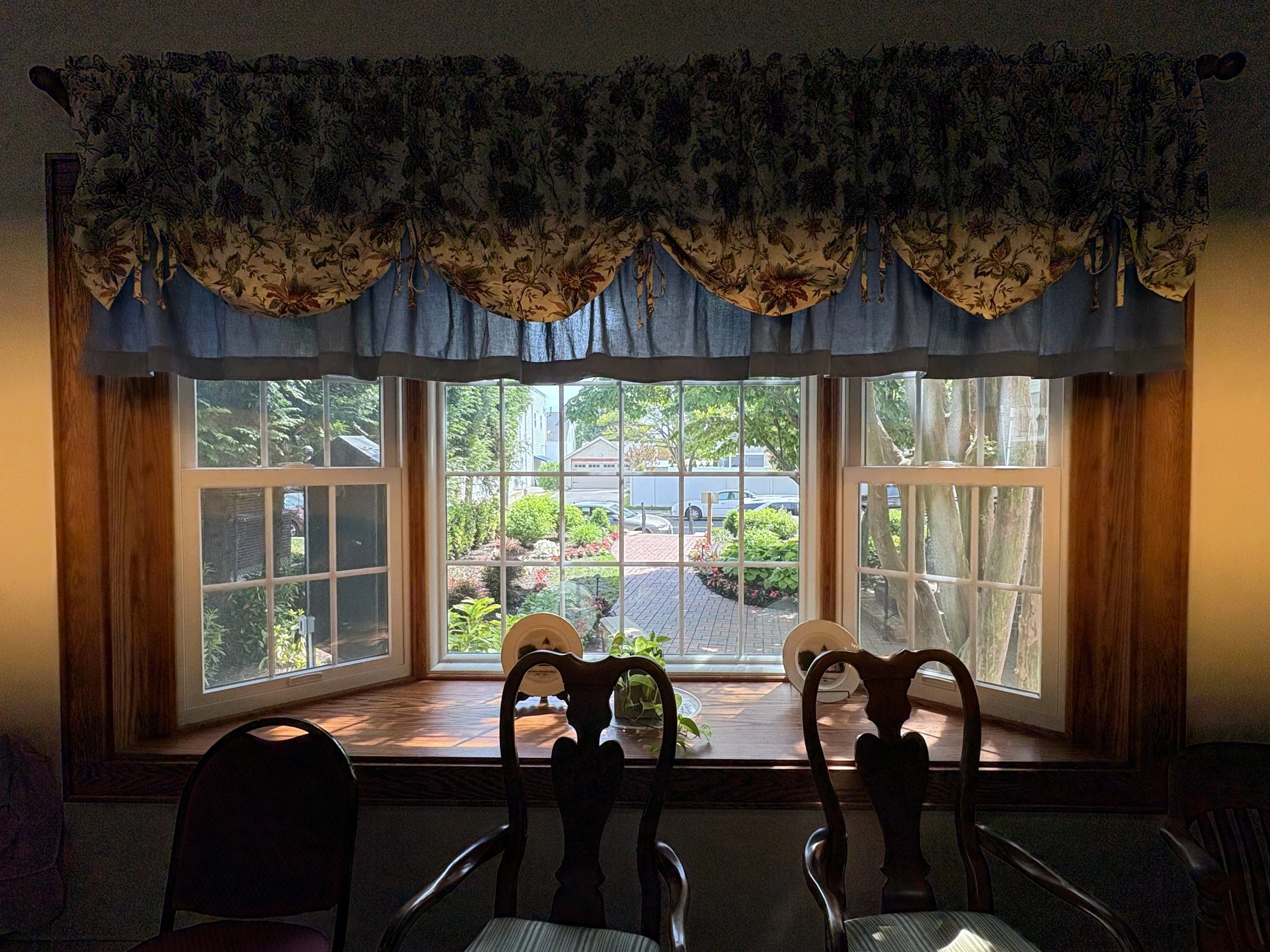A large window with a floral valance curtain and a sheer curtain behind it, overlooking a garden with trees, shrubs, and a brick pathway outside.