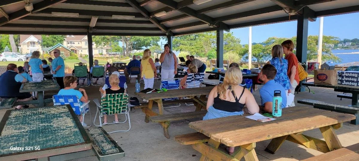 People gathered under a pavilion near a body of water, sitting at picnic tables and talking during daytime.