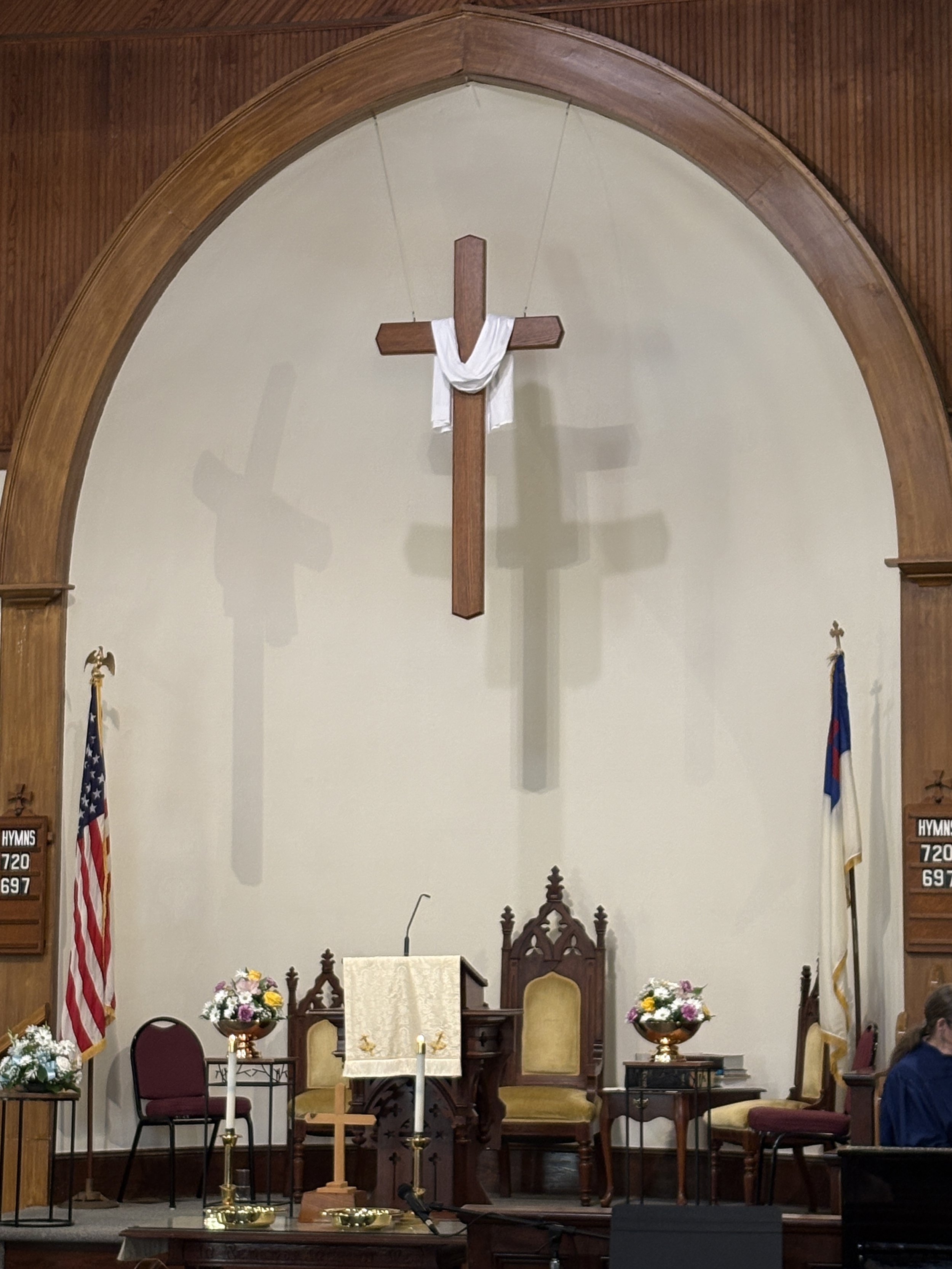 Interior view of a church altar with wooden cross draped in white cloth, surrounded by flowers, candles, and chairs, with flags on either side, and hymn boards displaying hymn numbers.