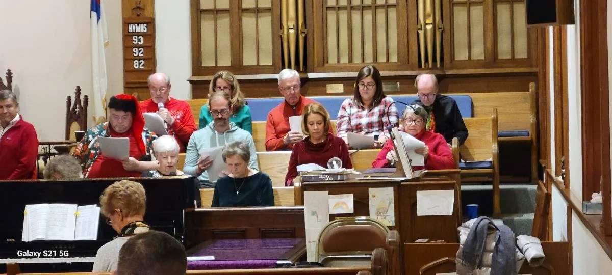 A group of people, mostly women, singing in a church choir, with some holding sheet music, seated on choir benches and at a piano, in a church with wooden decor and pipe organ pipes in the background.