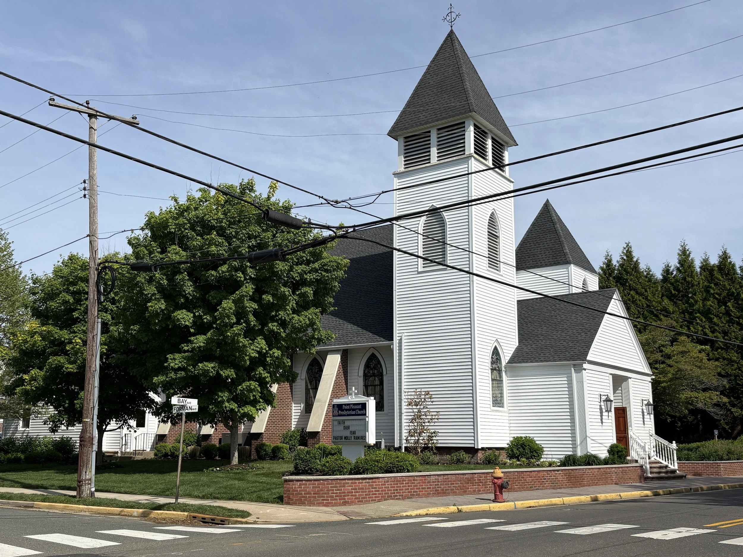 White church with a tall steeple and a dark roof, situated behind green trees and a brick wall, with street signs and power lines in the foreground.