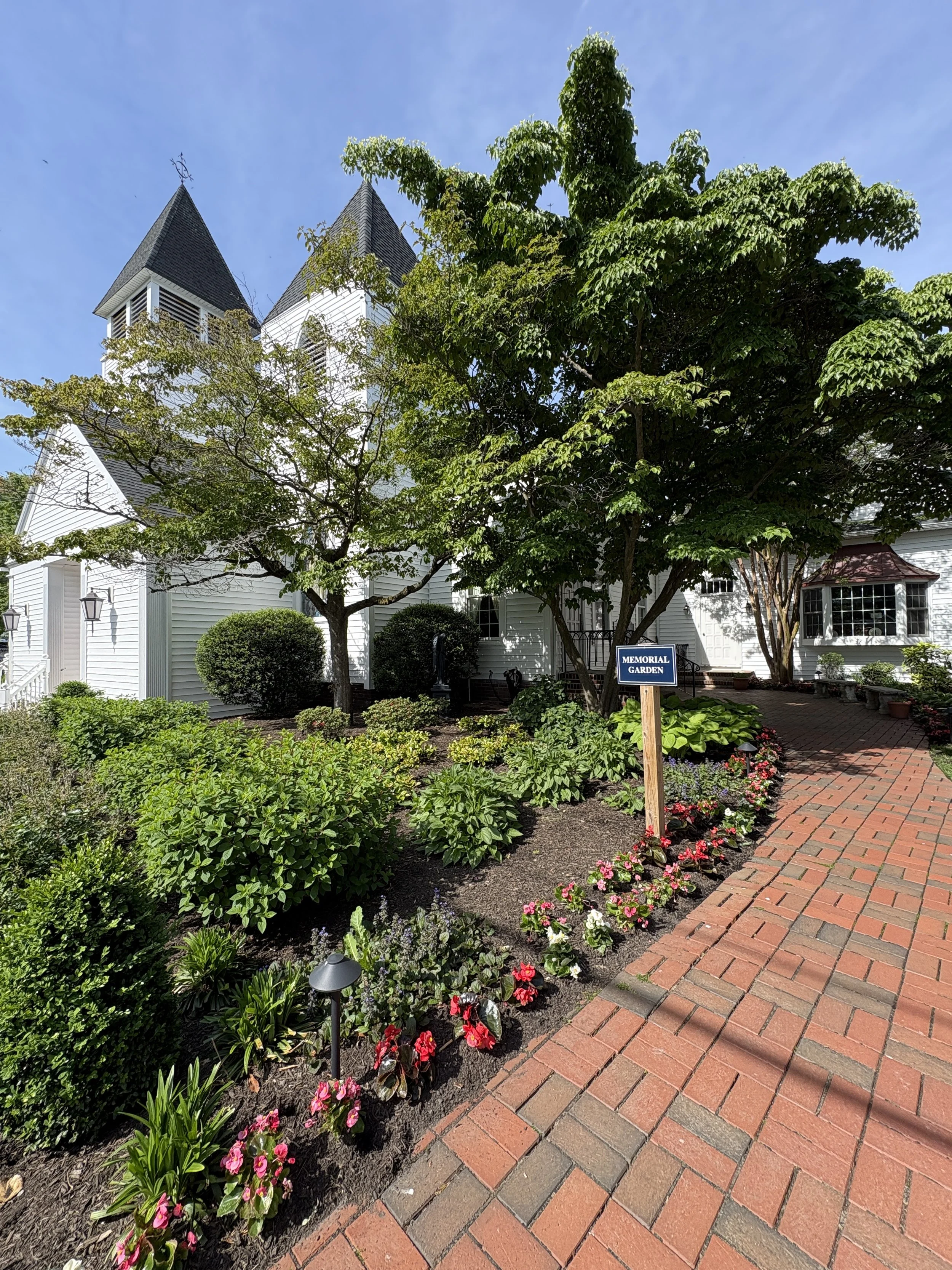 A church with two steeples next to a garden with flowers and plants. There is a sign that says 'MEMORIAL GARDEN' near the plants and a brick pathway.