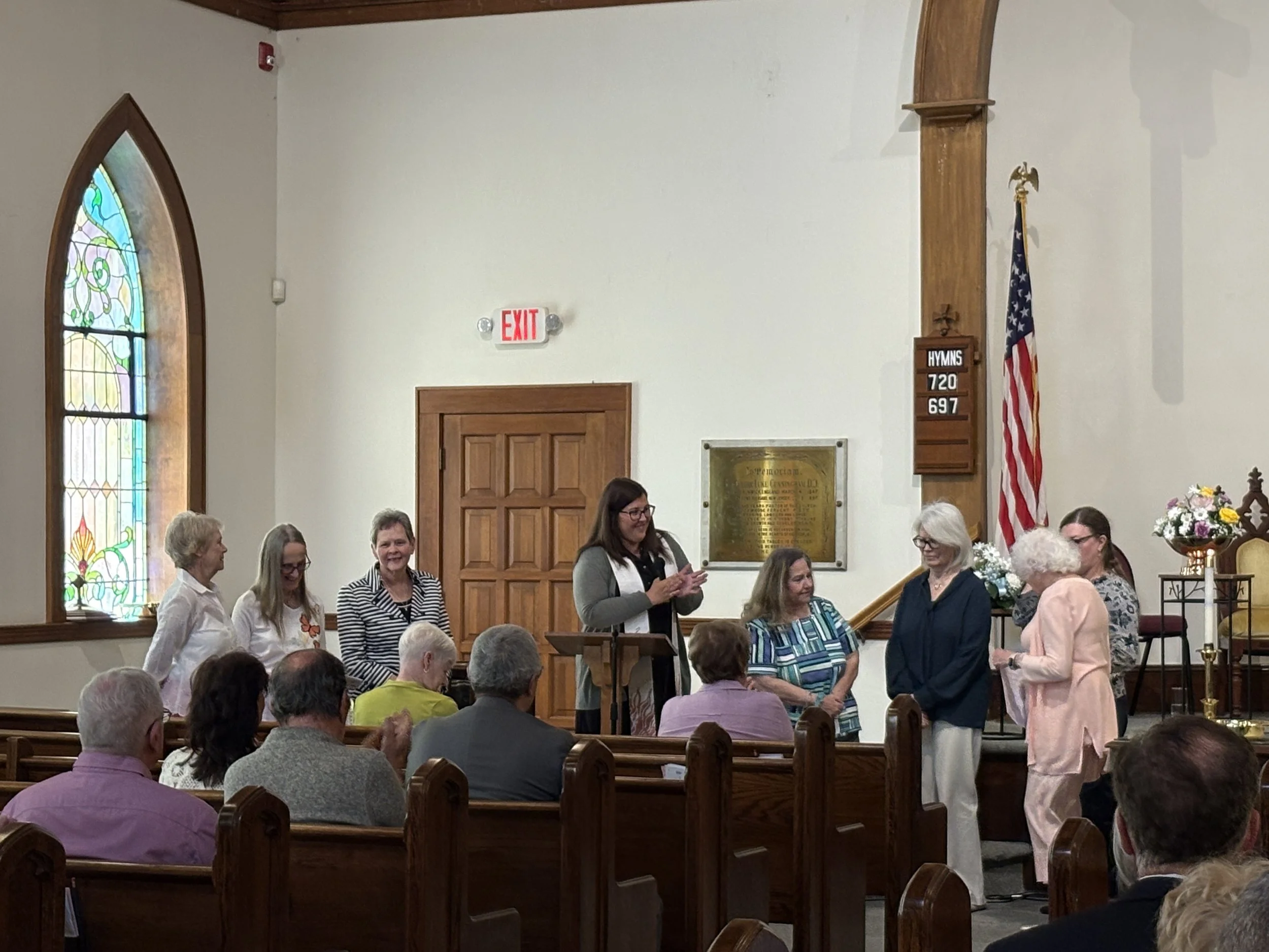 A group of women standing and interacting inside a church during a gathering, with people seated in pews, a stained glass window, a wooden door, a church plaque, and the American flag visible in the background.