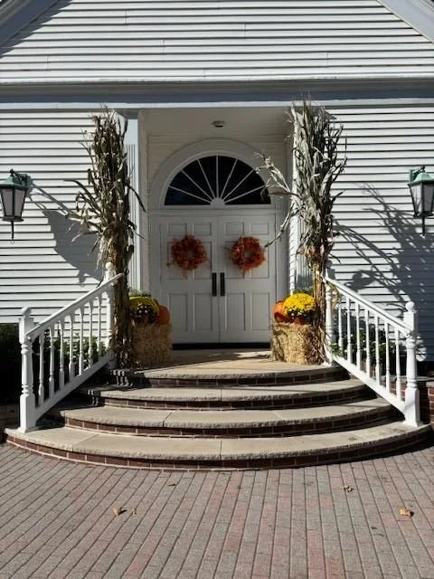 Front porch of a house with white double doors decorated with two orange wreaths. Cornstalks in baskets frame the doors. Two lantern-style lights are mounted on the house on either side of the porch. The steps are brick and curved, leading up to the 