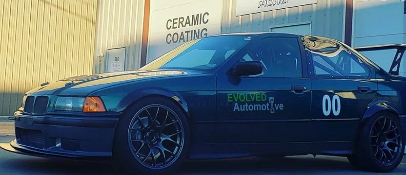A black race car with the number 00 on the side, parked outside a building with signs that read 'CERAMIC COATING'. The car has a large rear wing and aftermarket wheels.