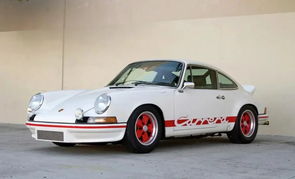 White Porsche 911 Carrera with red wheels and red stripe detail, parked against a beige wall.